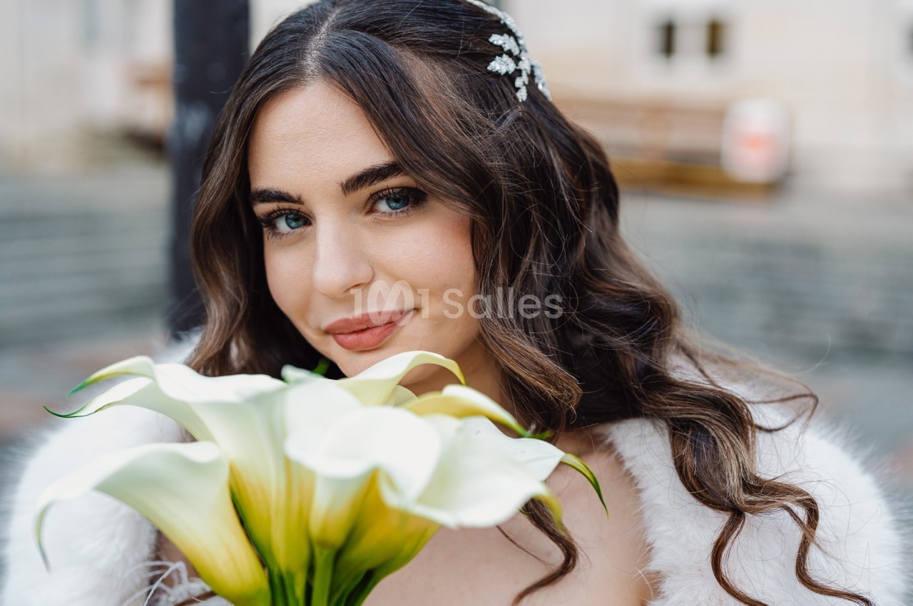 Femme souriante tenant un bouquet de lys blancs, cheveux bouclés ornés d'une barrette, en extérieur flou.