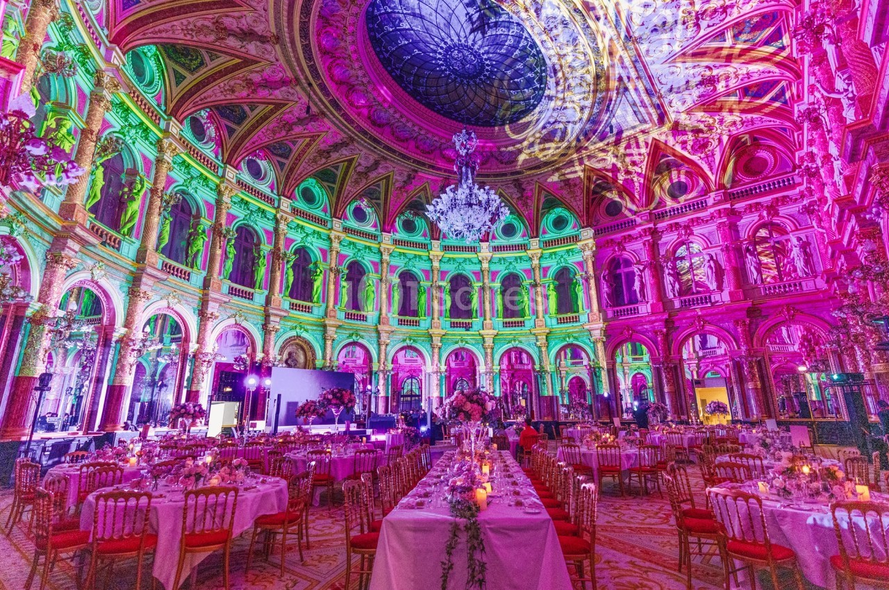 Salle de réception luxueuse avec plafond orné, éclairage coloré et tables dressées pour un événement.