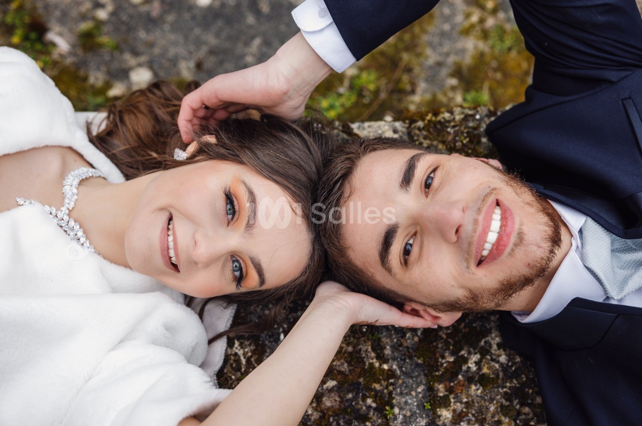 Un couple allongé côte à côte, souriant, vêtu élégamment, avec un fond de pierre et de végétation.