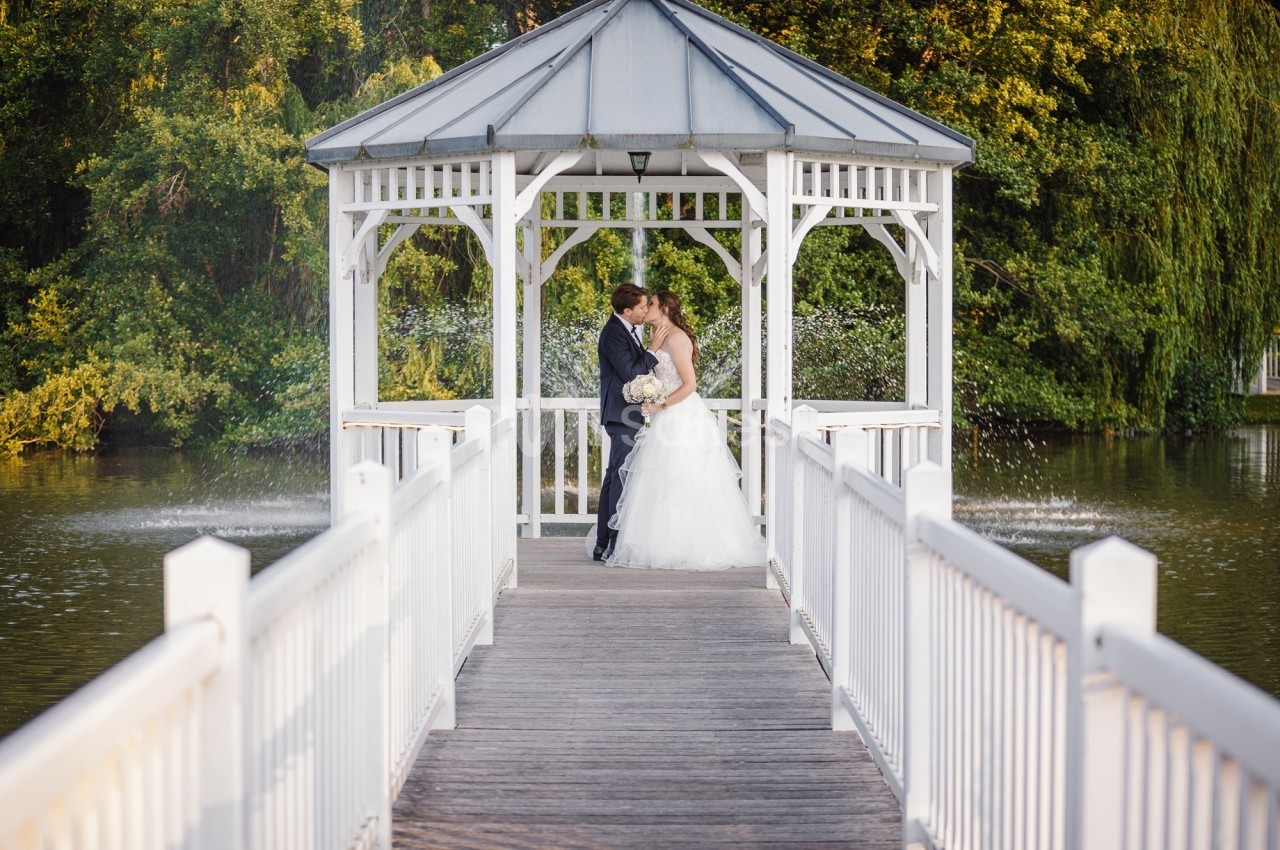 Un couple de mariés s'embrasse sous une pergola blanche au bord d'un lac, entourée de végétation.
