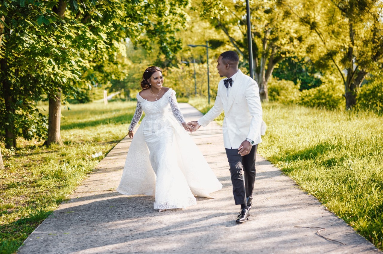 Un couple en tenue de mariage marche main dans la main sur un chemin entouré de verdure.