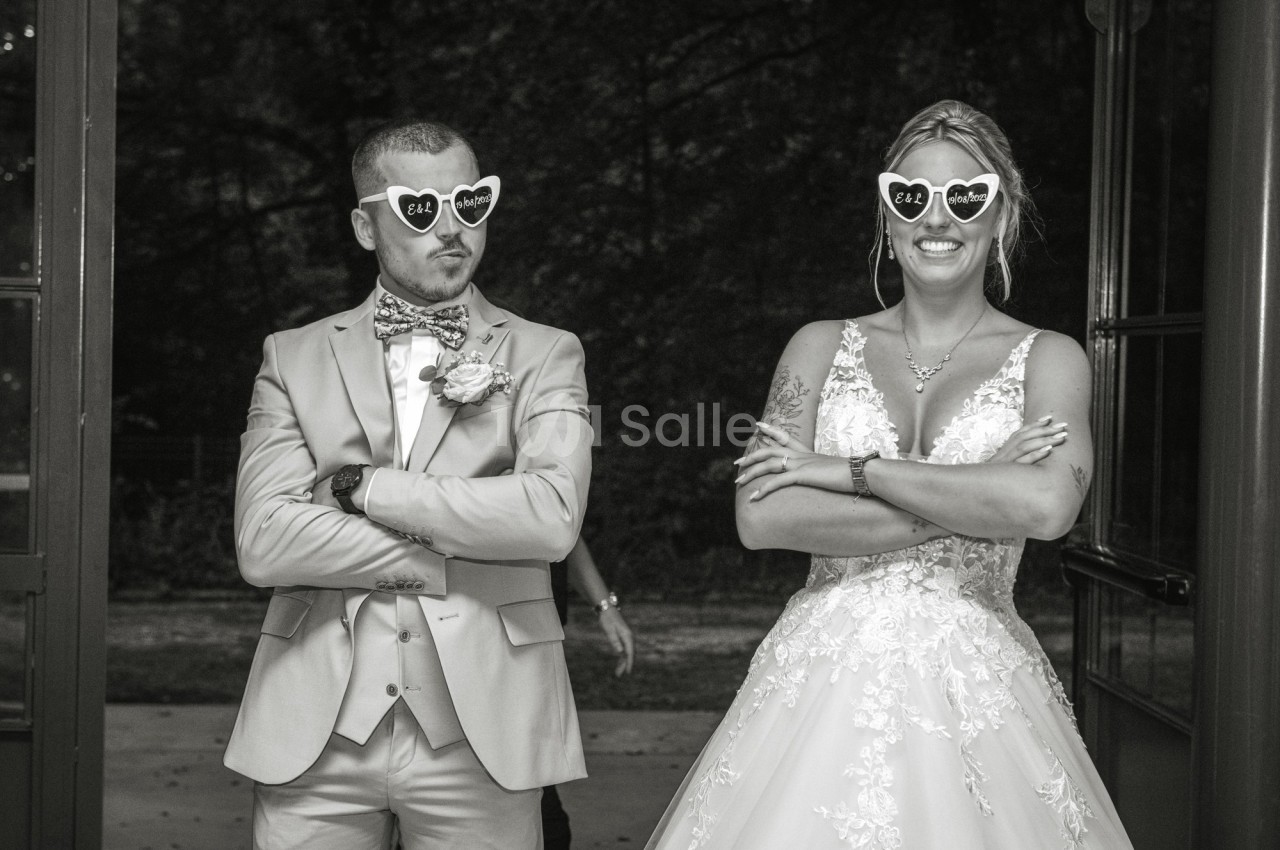 Un couple en tenue de mariage pose avec des lunettes fantaisie en forme de cœur, bras croisés et sourires amusés.