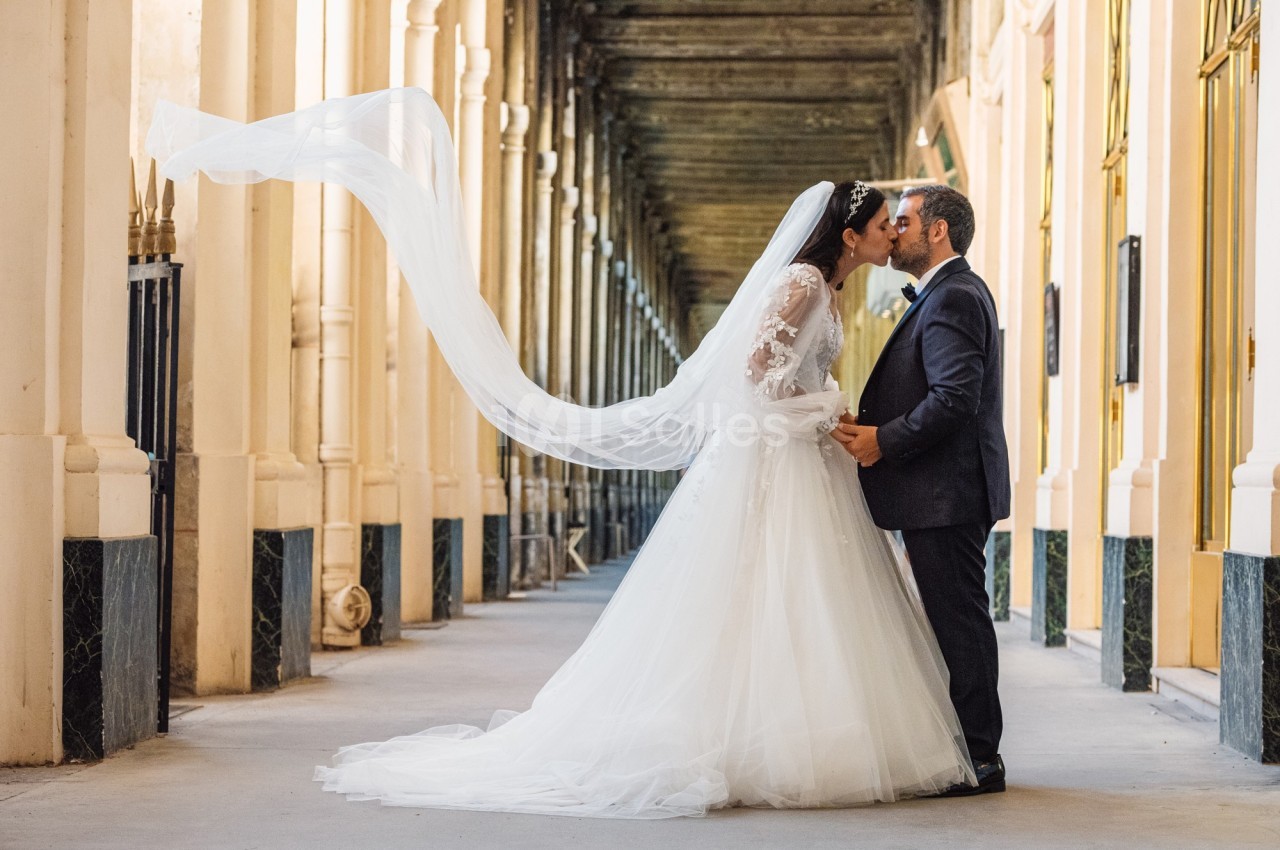Un couple de mariés s'embrasse dans une galerie avec une architecture élégante, le voile de la mariée flottant au vent.