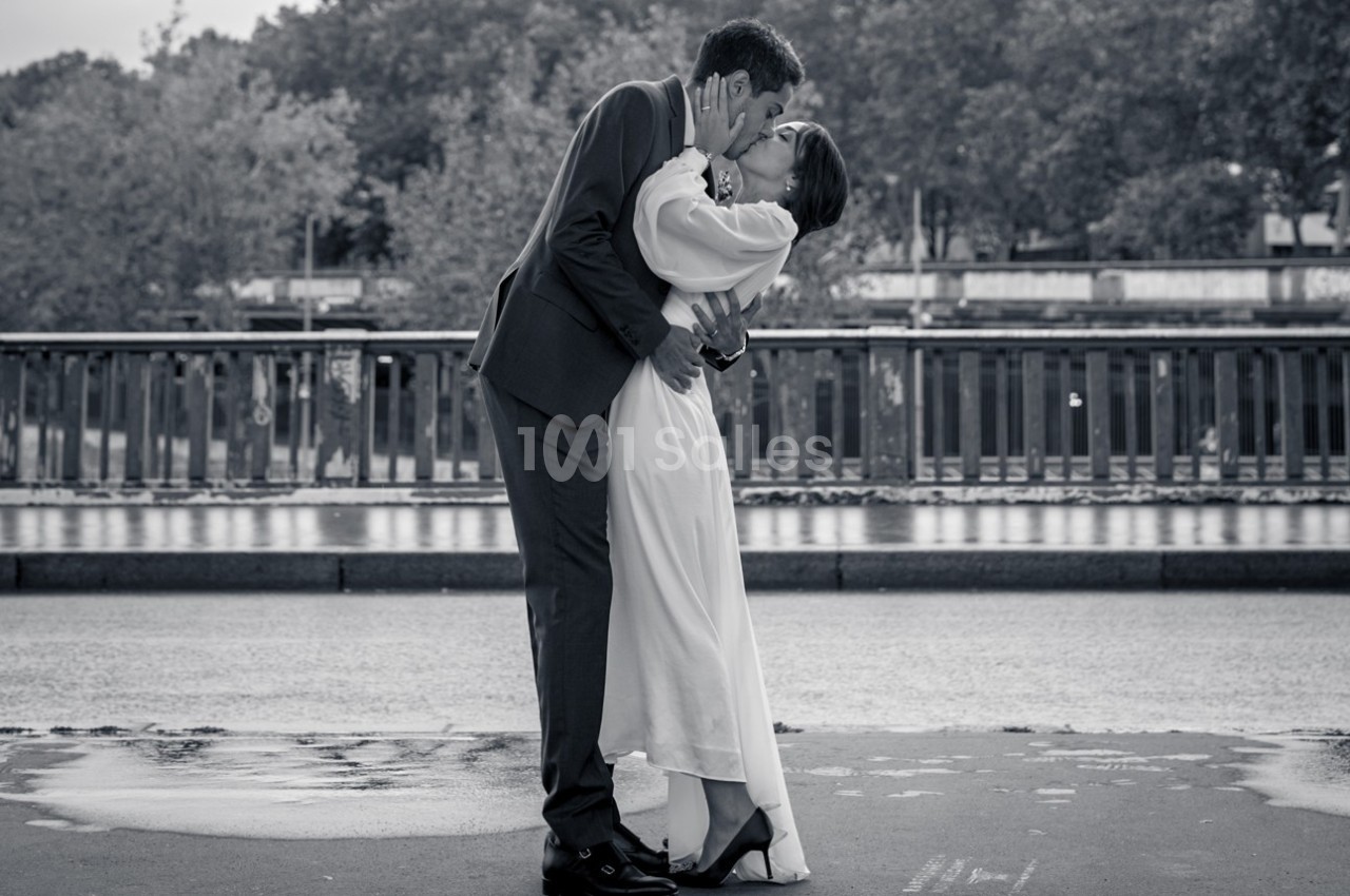 Un couple en tenue de mariage s'embrasse tendrement sur un pont, avec des arbres en arrière-plan.