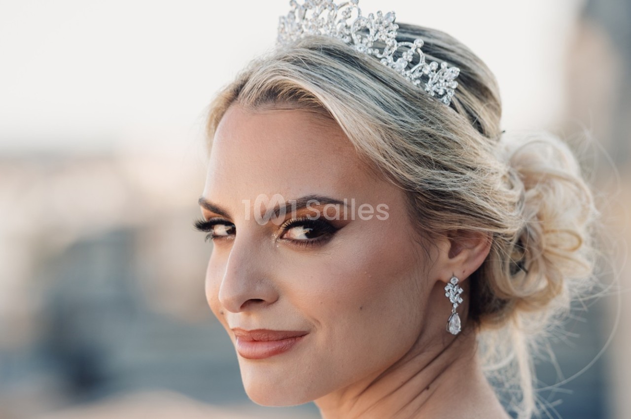 Femme blonde portant une couronne ornée de pierres et des boucles d'oreilles, coiffée d'un chignon élégant.