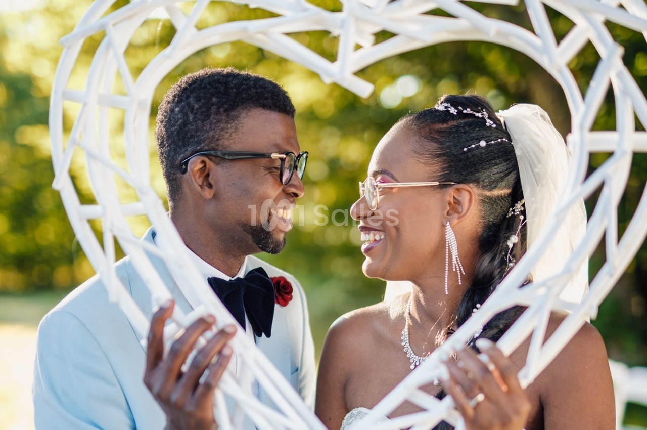 Un couple souriant se regarde à travers un cadre en forme de cœur, dans un décor extérieur ensoleillé.
