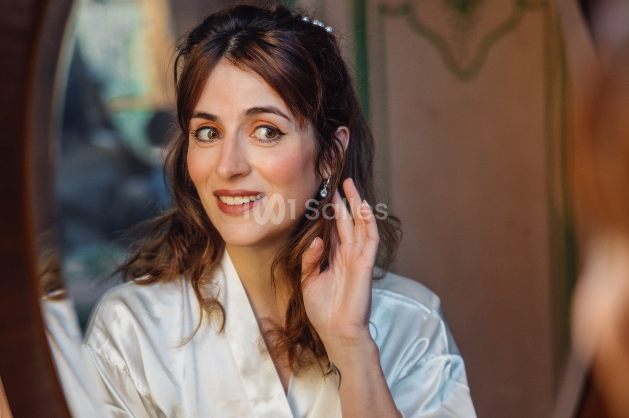 Femme en robe de satin blanche se regardant dans un miroir tout en ajustant une boucle d'oreille.