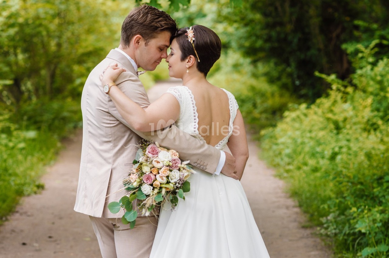 Un couple de mariés s'enlace tendrement sur un chemin bordé de verdure, la mariée tenant un bouquet de fleurs pastel.