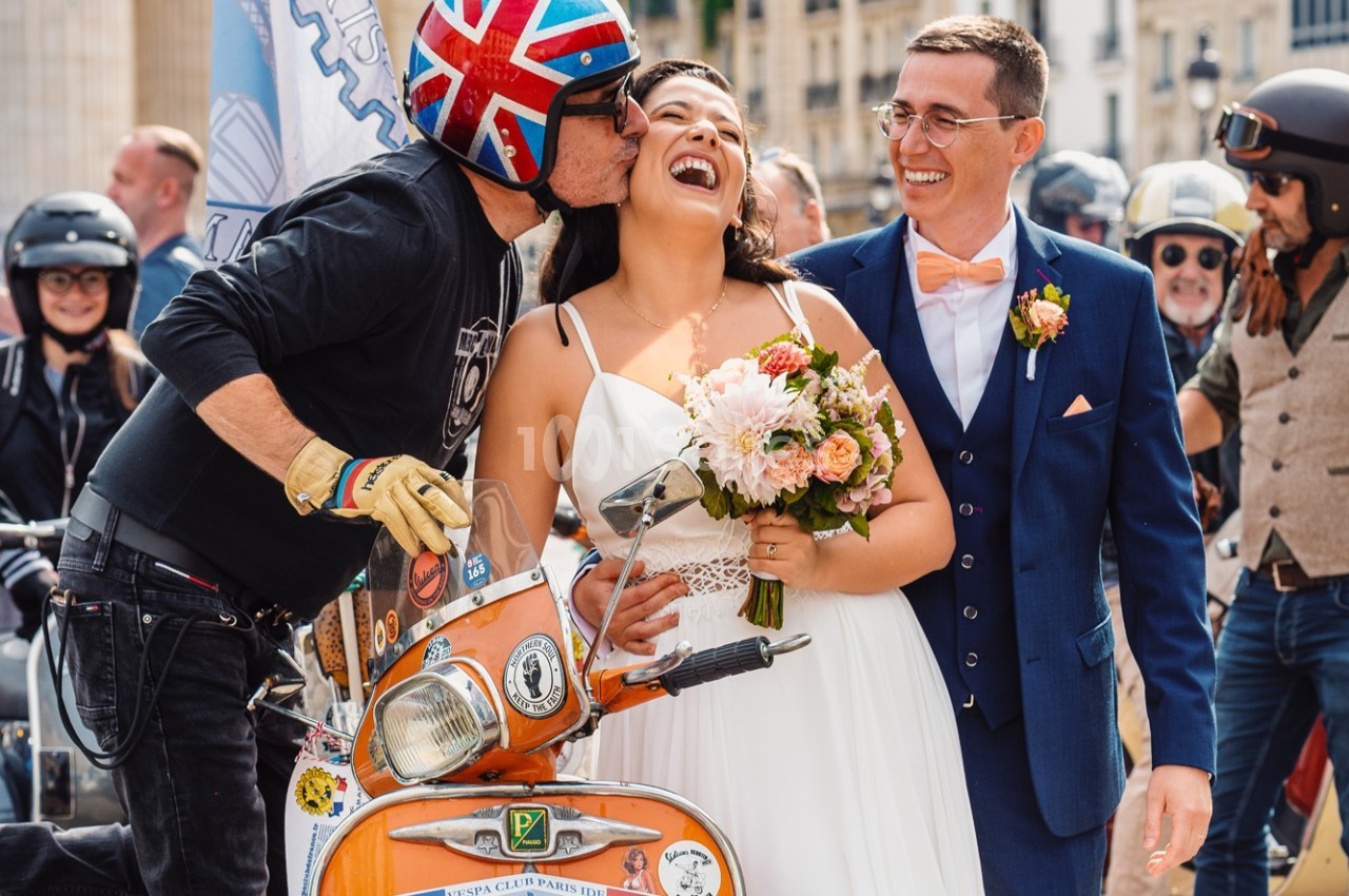 Un couple de mariés souriant, entouré d'un motard embrassant la mariée près d'un scooter orange.