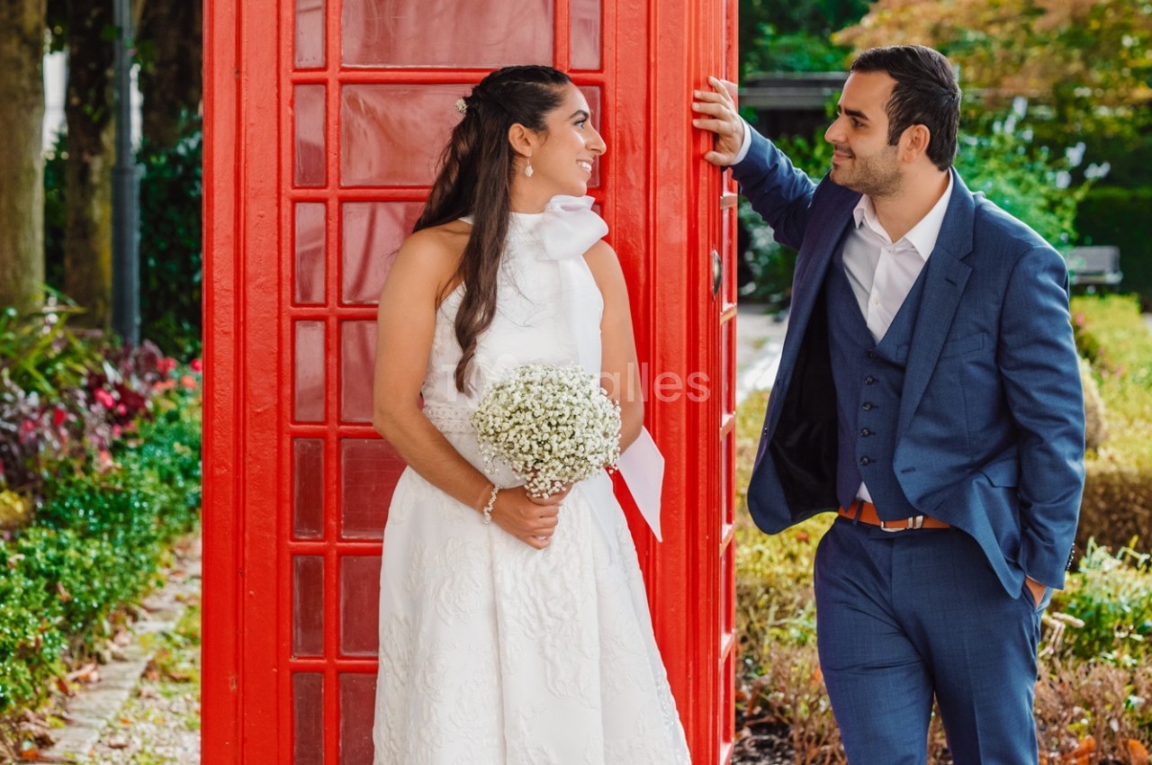 Une mariée en robe blanche et un homme en costume discutent près d'une cabine téléphonique rouge dans un parc.