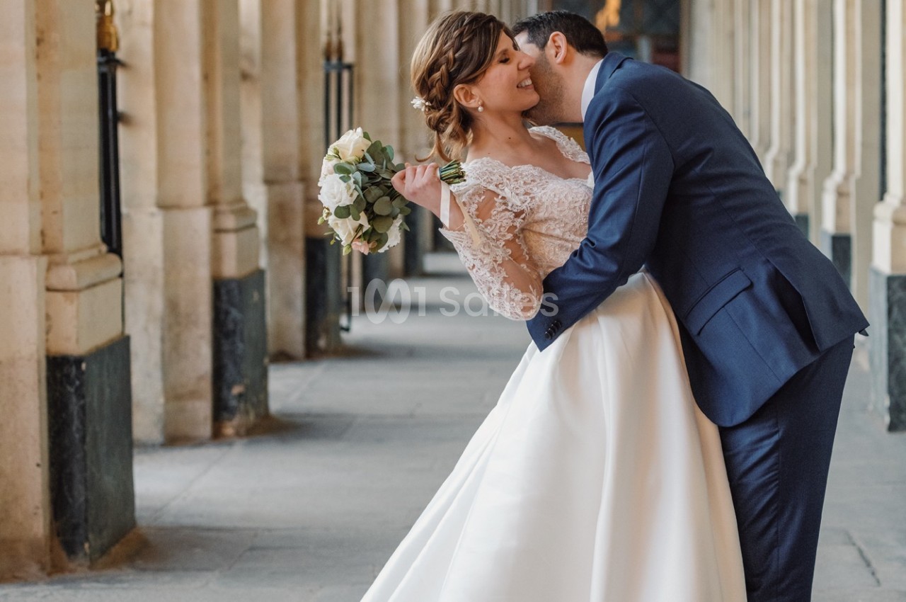 Un couple de mariés s'embrasse dans une galerie avec des colonnes, la mariée tenant un bouquet de fleurs.