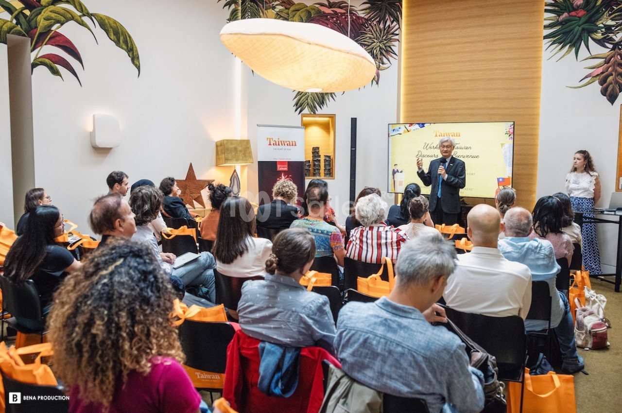Un conférencier s'adresse à un public assis dans une salle décorée de motifs tropicaux.