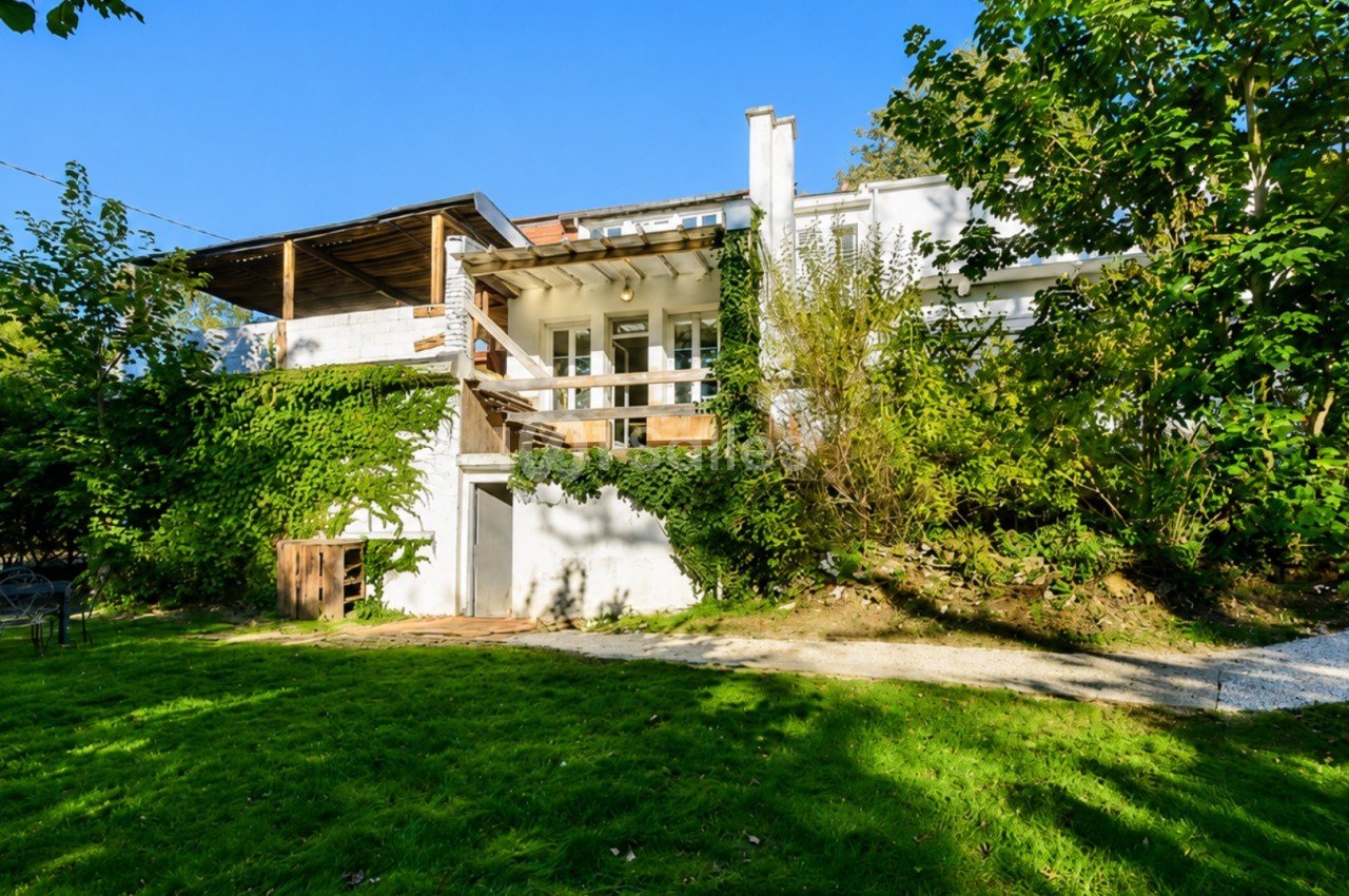 Façade d'une maison blanche entourée de verdure, avec terrasse en bois et jardin ensoleillé.