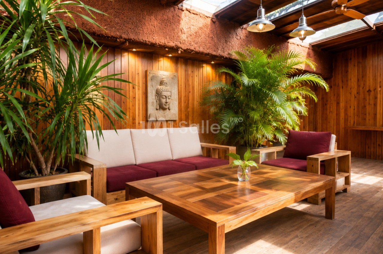 Espace salon chaleureux avec mobilier en bois, coussins rouges, plantes vertes et lumière naturelle entrant par le toit.
