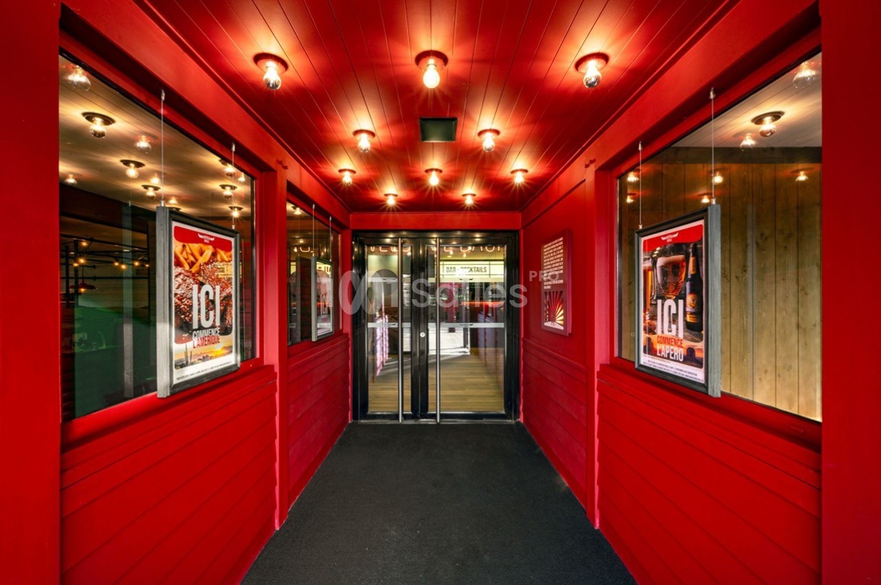 Entrée d'un bâtiment avec murs rouges, affiches publicitaires et portes vitrées au fond, éclairée par des lampes au plafond.