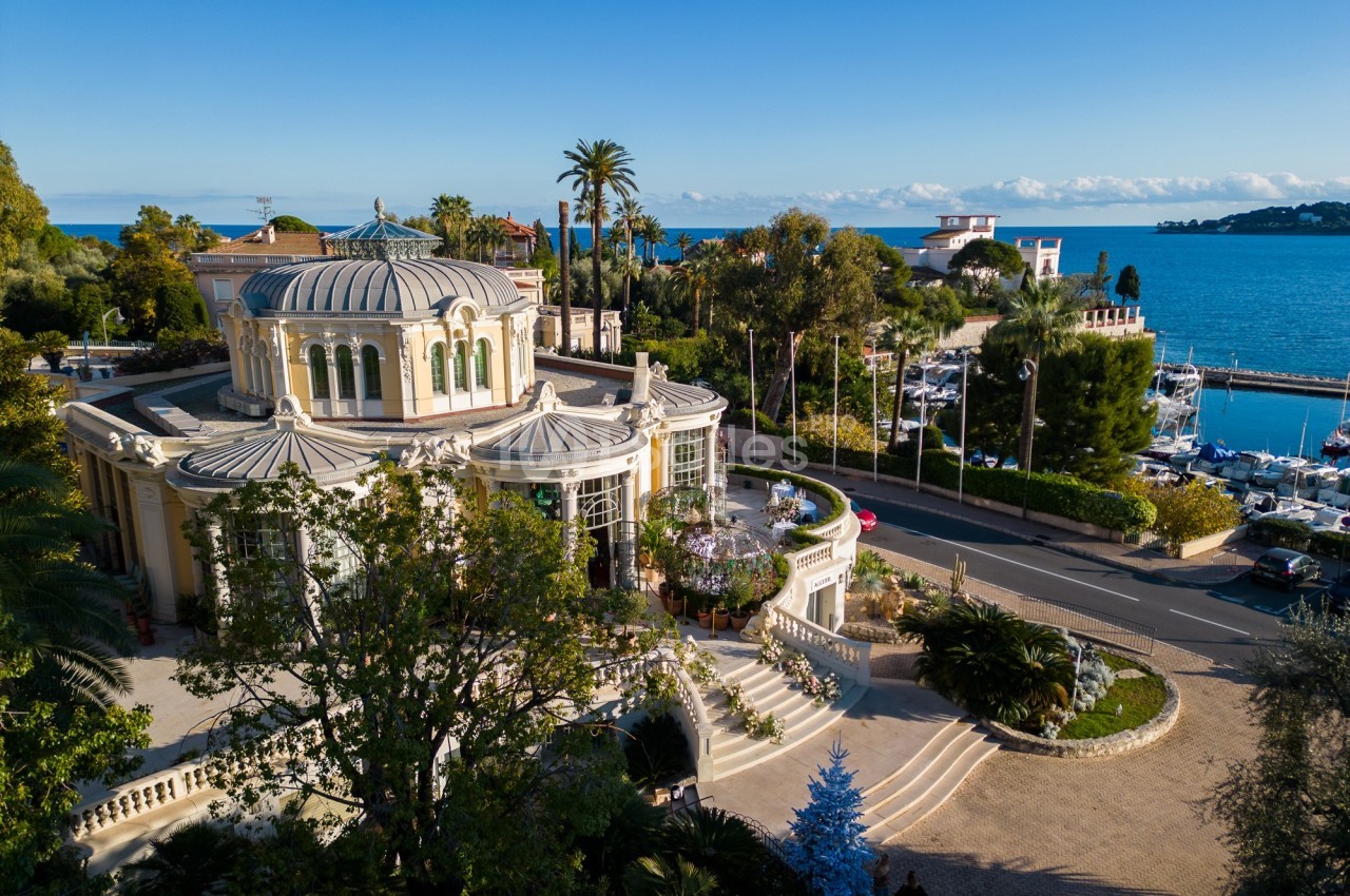 Vue aérienne d'un bâtiment élégant entouré de végétation, près d'une route, avec la mer et un port en arrière-plan.