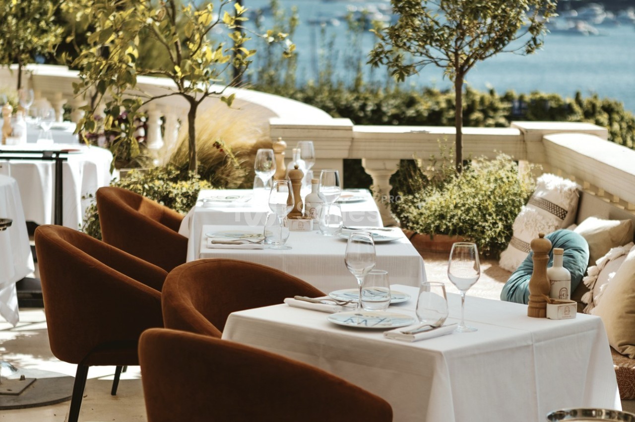 Terrasse de restaurant élégante avec tables dressées, chaises confortables et vue sur un paysage côtier en arrière-plan.