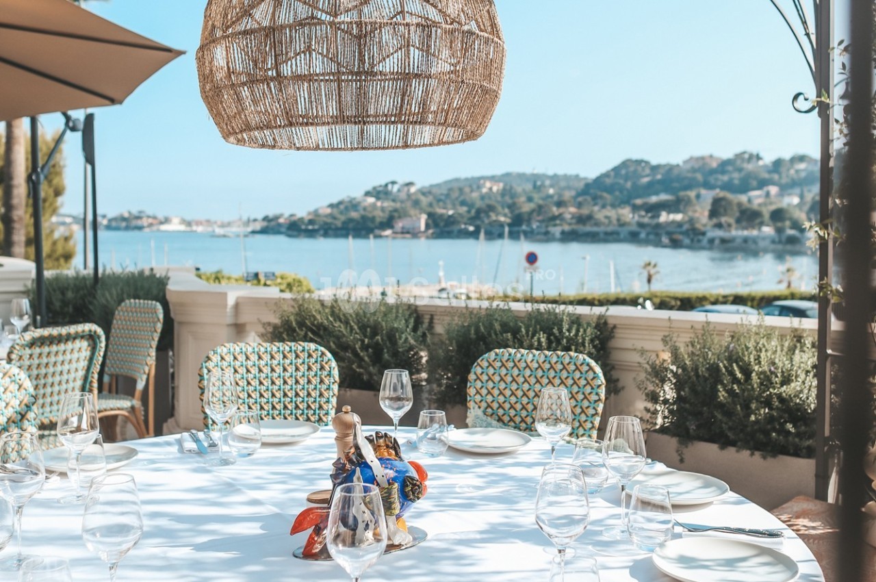 Table dressée en extérieur avec vue sur une baie, des collines et des bateaux, sous une suspension en osier.