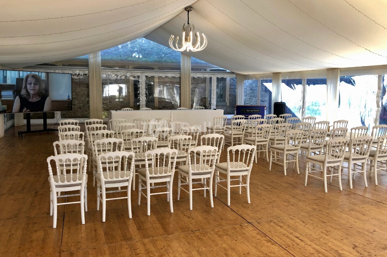 Salle de réception avec des chaises blanches alignées face à une table, sous une tente avec un lustre central.