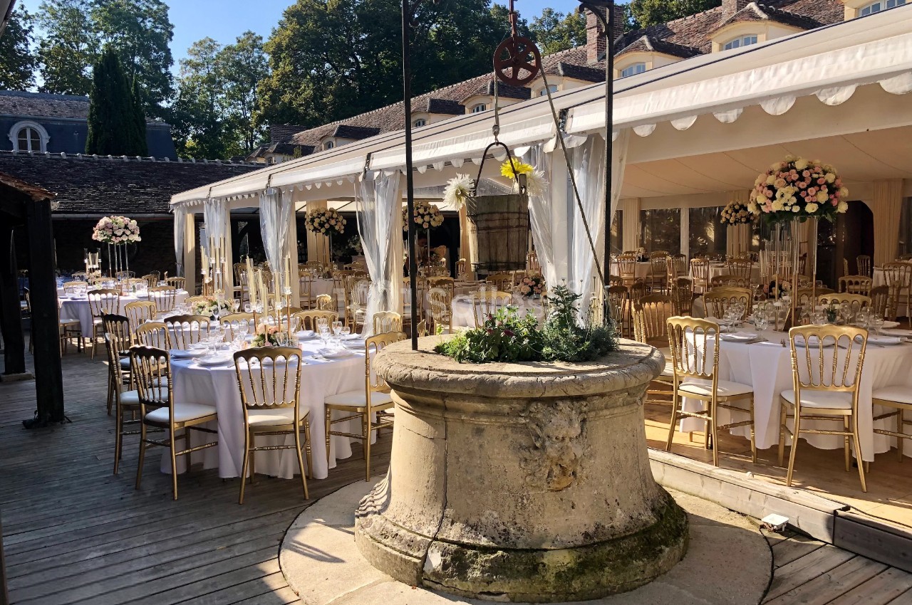 Tables élégamment dressées sous une tente blanche, entourées de chaises dorées, avec un puits central décoré de fleurs.