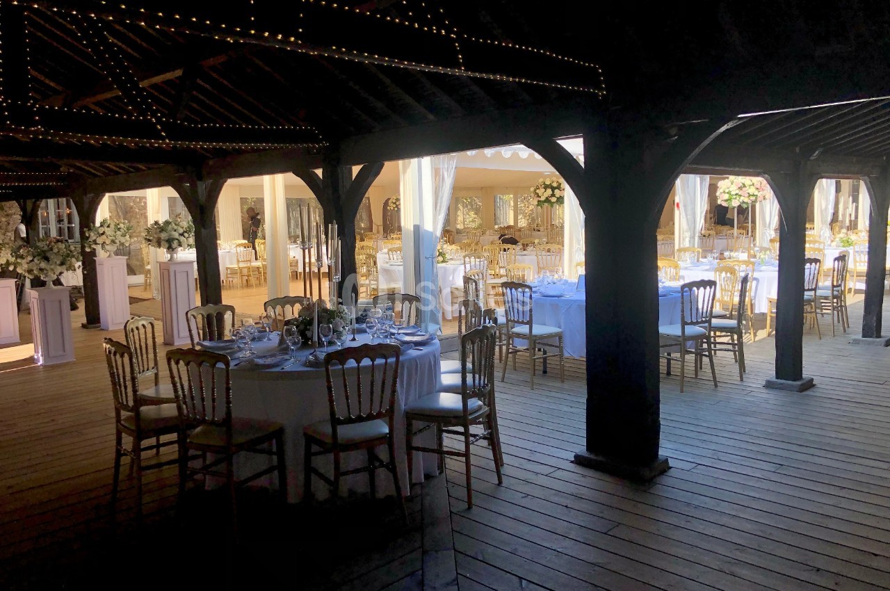 Salle de réception en bois avec tables rondes dressées pour un événement, éclairée par des guirlandes lumineuses.