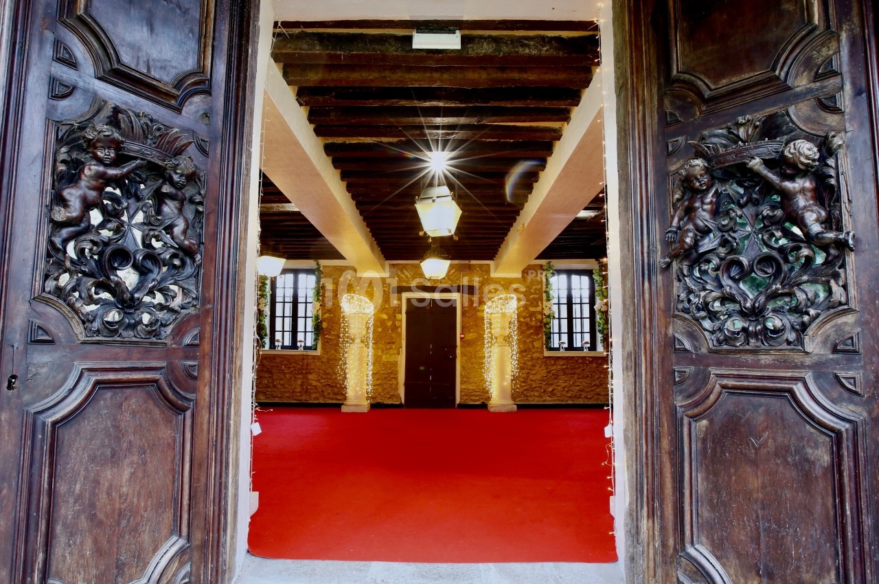 Vue intérieure d'une salle avec un sol rouge, des murs en pierre, des poutres apparentes et des portes en bois sculpté.