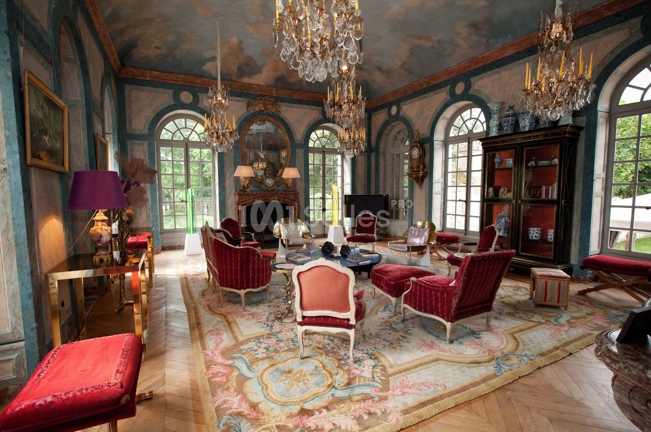 Salon élégant avec mobilier classique, lustres en cristal, murs peints et grandes fenêtres donnant sur un jardin.