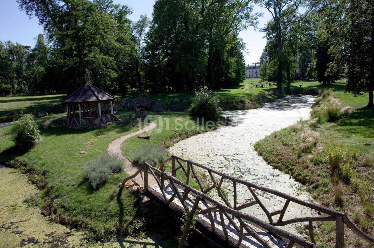 Un pont en bois traverse un ruisseau bordé de végétation dans un parc avec un kiosque et un bâtiment en arrière-plan.