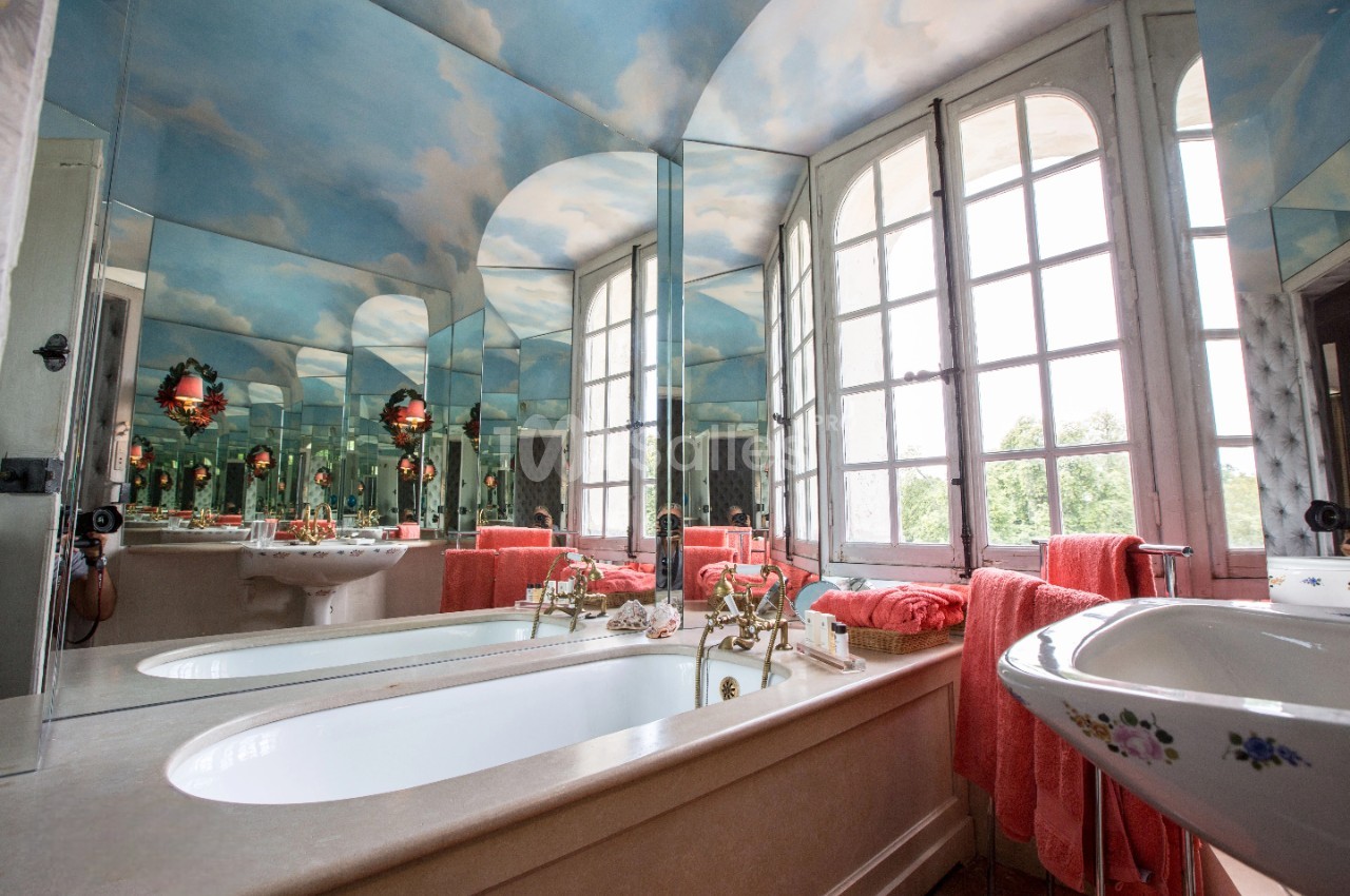 Salle de bain lumineuse avec baignoire, lavabo fleuri, miroirs, serviettes rouges et plafond peint en ciel nuageux.