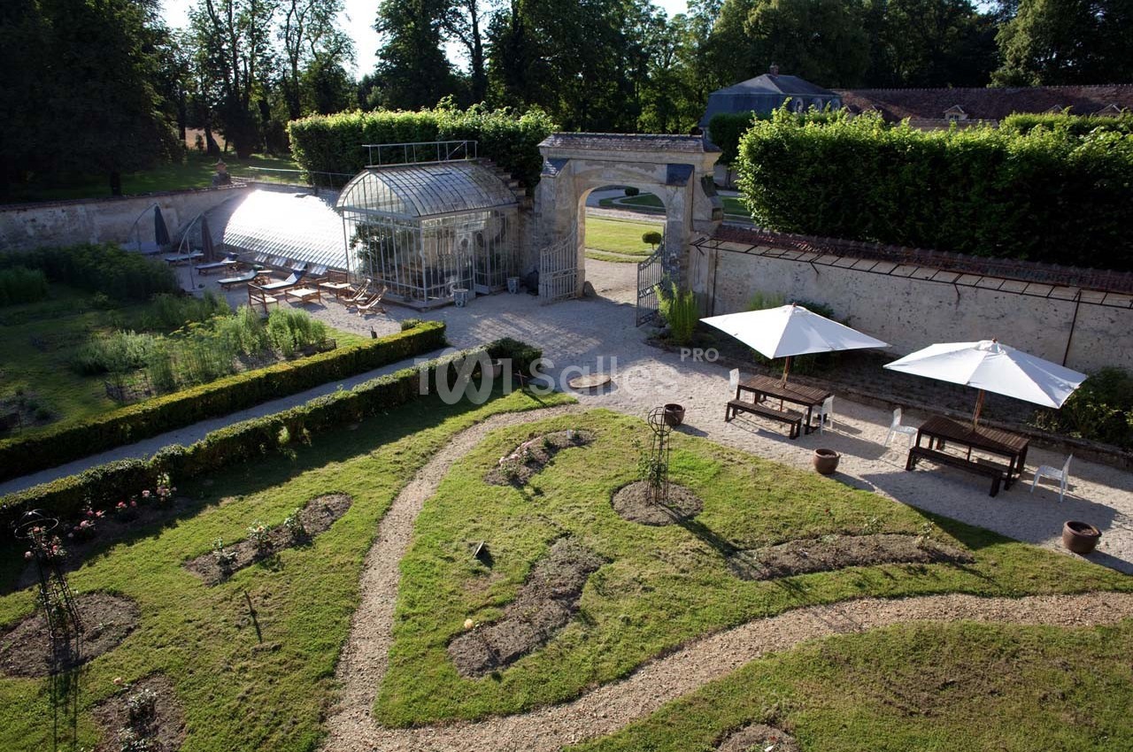 Jardin paysager avec allées sinueuses, parterres de plantes, serre en verre et bancs sous des parasols blancs.
