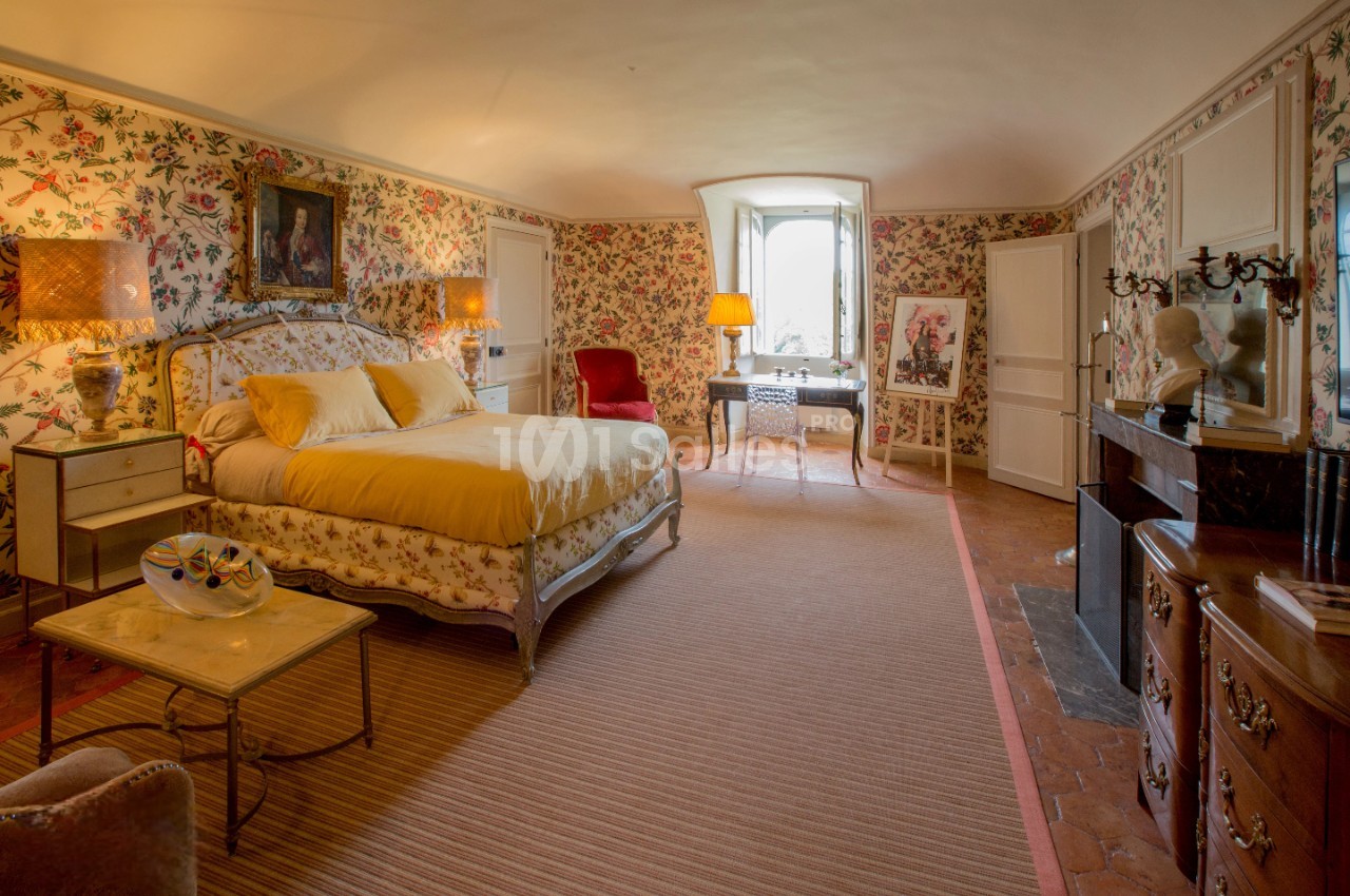 Chambre spacieuse avec lit double, mobilier ancien, cheminée, murs ornés de papier peint fleuri et lumière naturelle.
