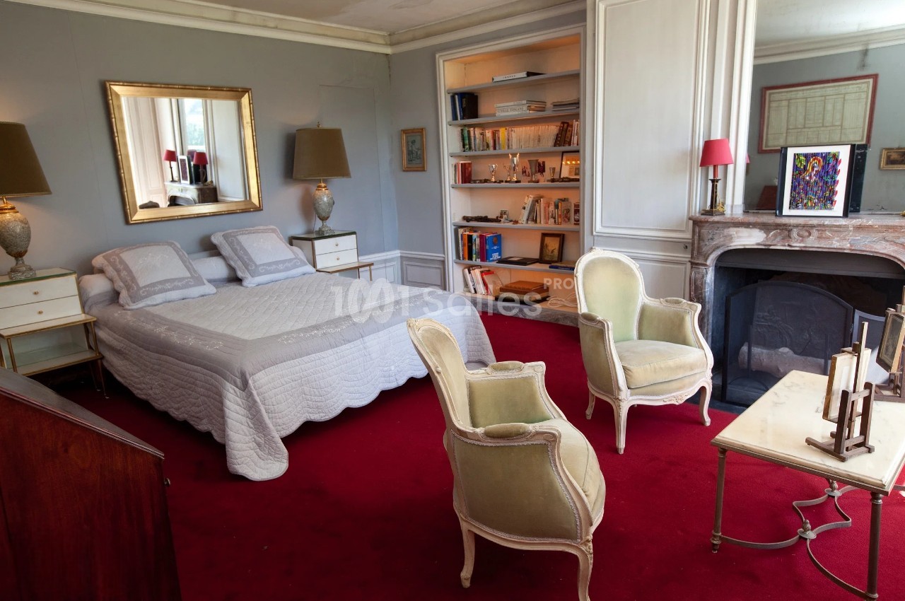 Chambre avec lit double, fauteuils anciens, cheminée, étagères remplies de livres et tapis rouge au sol.