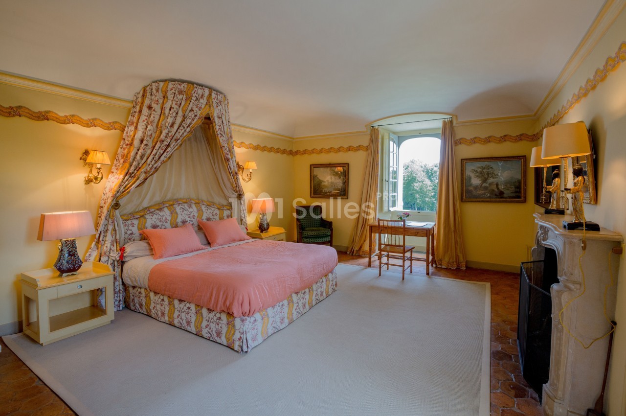 Chambre lumineuse avec lit à baldaquin, murs jaunes, mobilier classique et fenêtre donnant sur un paysage verdoyant.