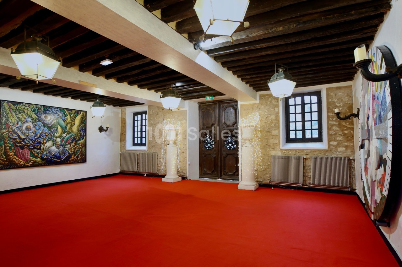 Salle avec un sol rouge, murs en pierre, poutres en bois au plafond, œuvres d'art accrochées et colonnes décoratives.
