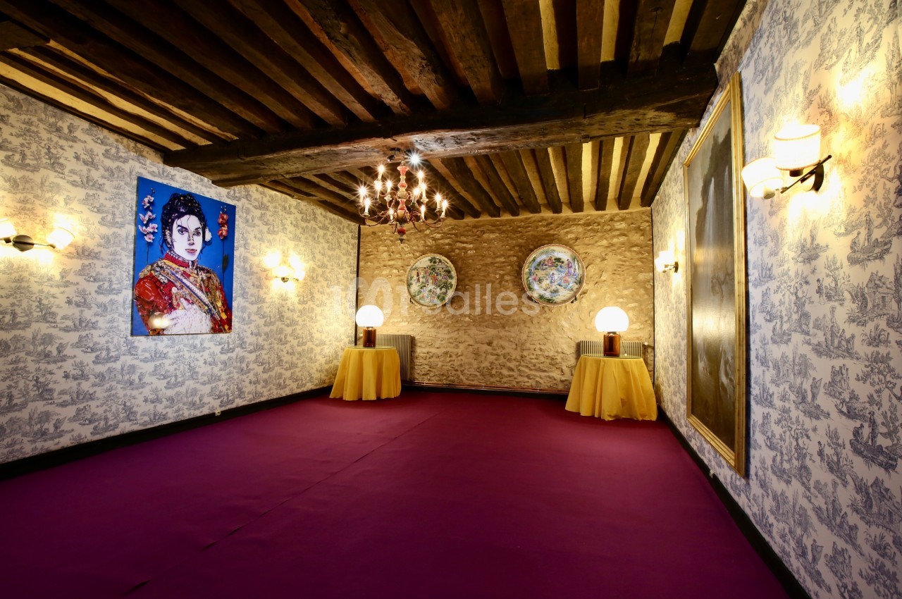 Salle décorée avec des murs en pierre et papier peint, plafond à poutres apparentes, tableaux et luminaires élégants.