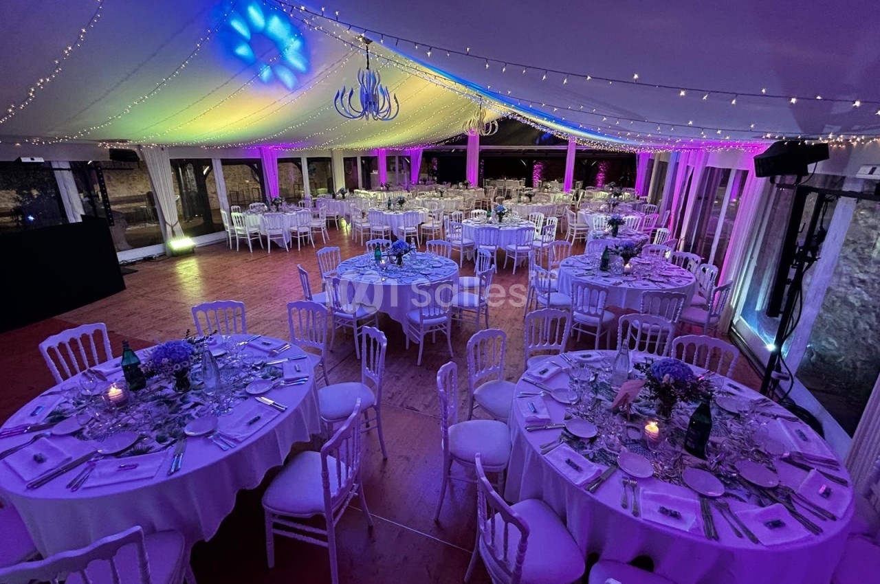 Salle de réception décorée avec des tables rondes dressées, éclairage coloré et guirlandes lumineuses au plafond.