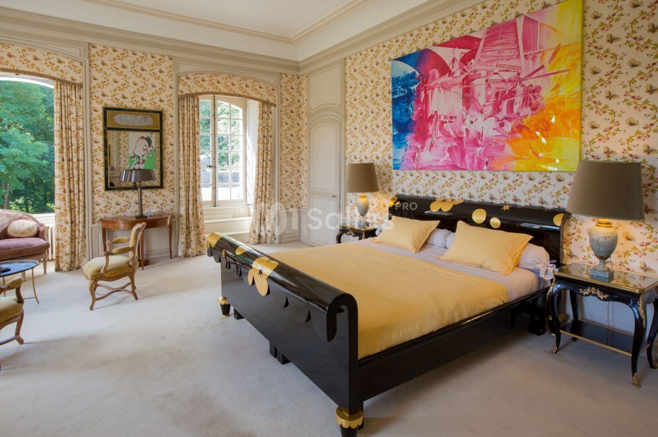 Chambre lumineuse avec lit double, mobilier classique, murs tapissés de motifs floraux et grande fenêtre donnant sur un…