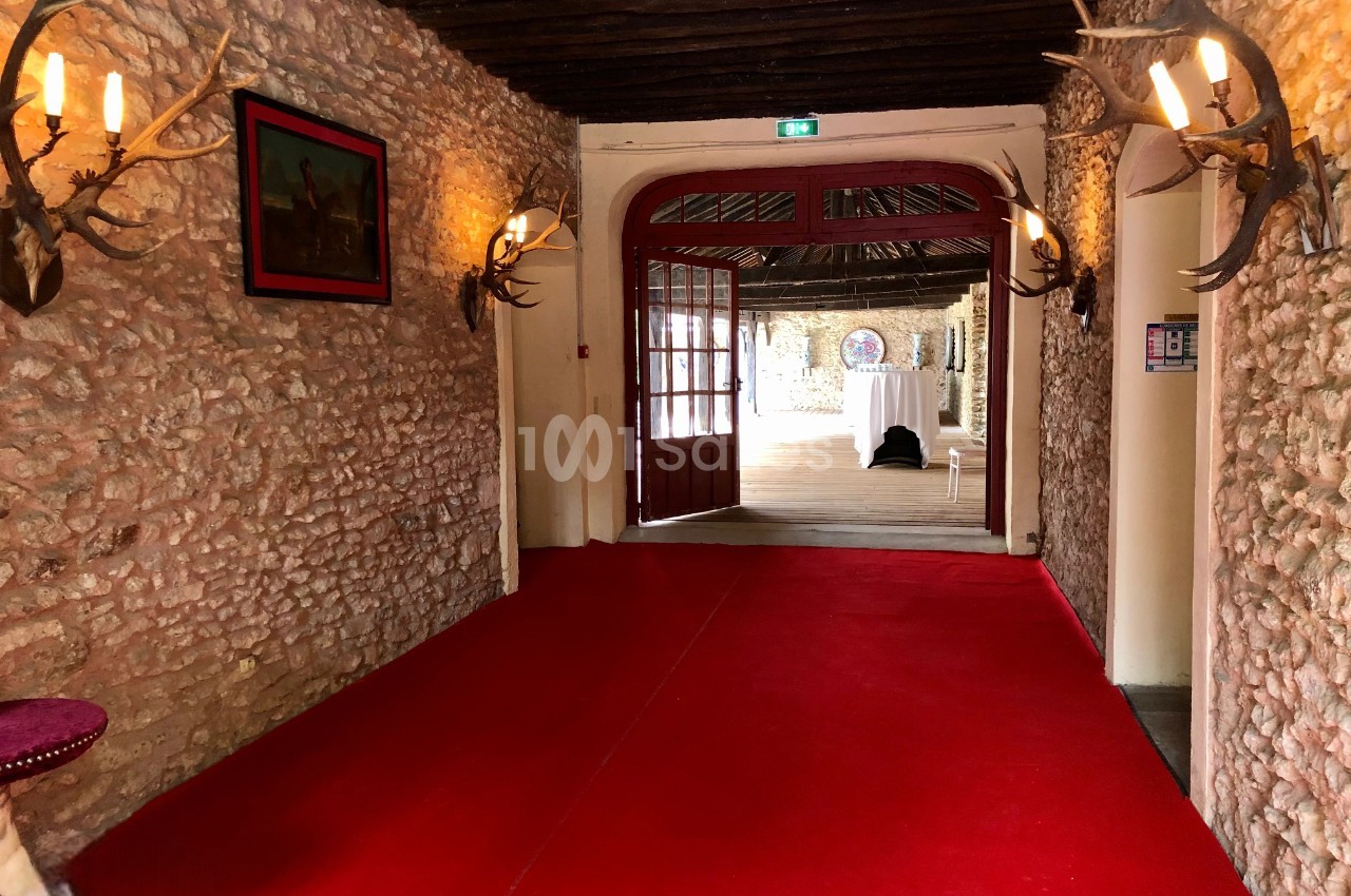 Couloir en pierre avec tapis rouge, éclairé par des appliques murales, menant à une porte ouverte sur une salle lumineuse.