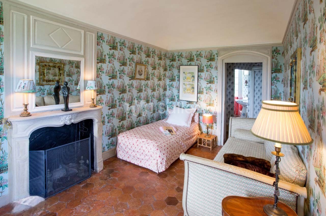 Chambre lumineuse avec lit simple, canapé, cheminée décorative et murs recouverts de papier peint fleuri.