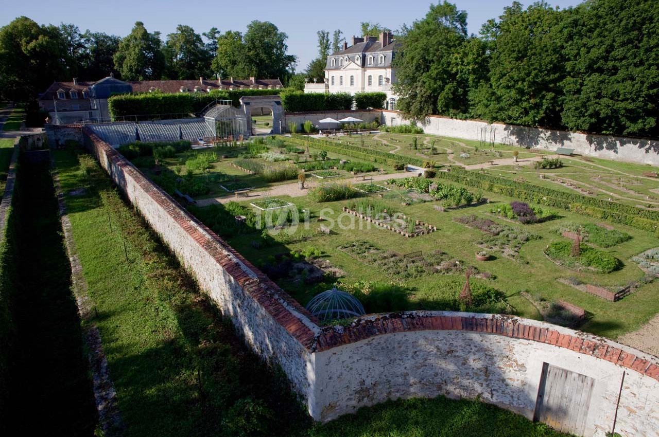 Jardin potager clos de murs avec allées symétriques, serres et un bâtiment historique en arrière-plan.