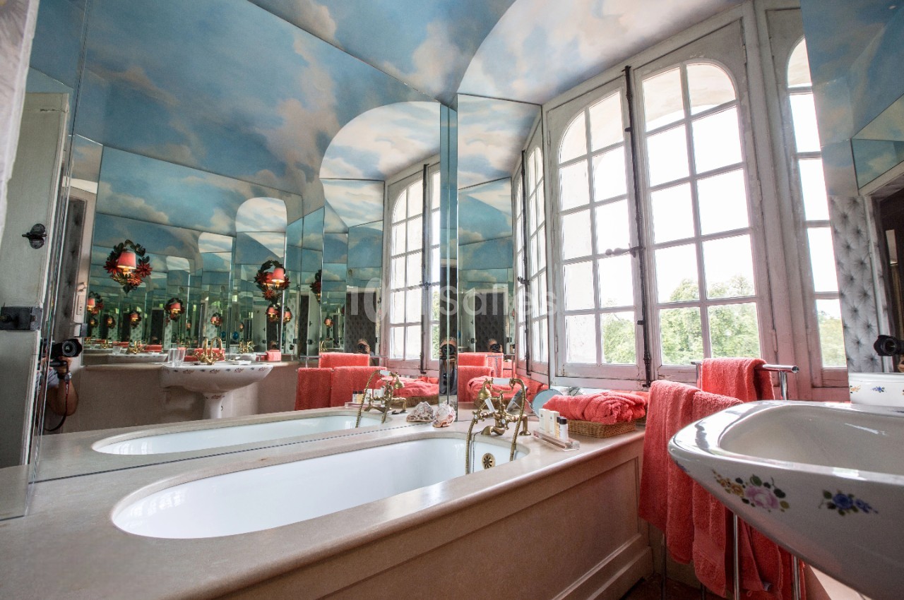 Salle de bain lumineuse avec baignoire, lavabo fleuri, miroirs, serviettes rouges et plafond peint en ciel nuageux.
