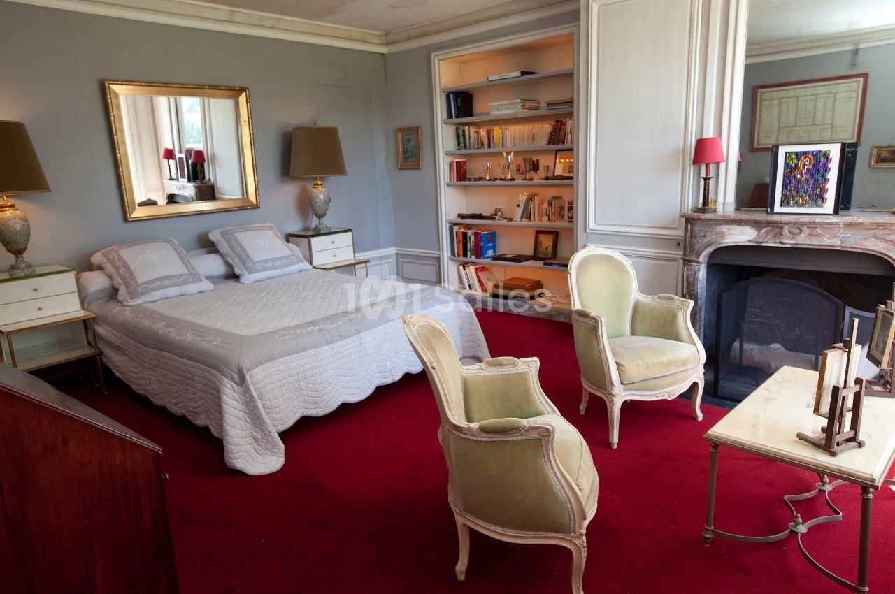 Chambre avec lit double, fauteuils anciens, cheminée, étagères remplies de livres et tapis rouge au sol.