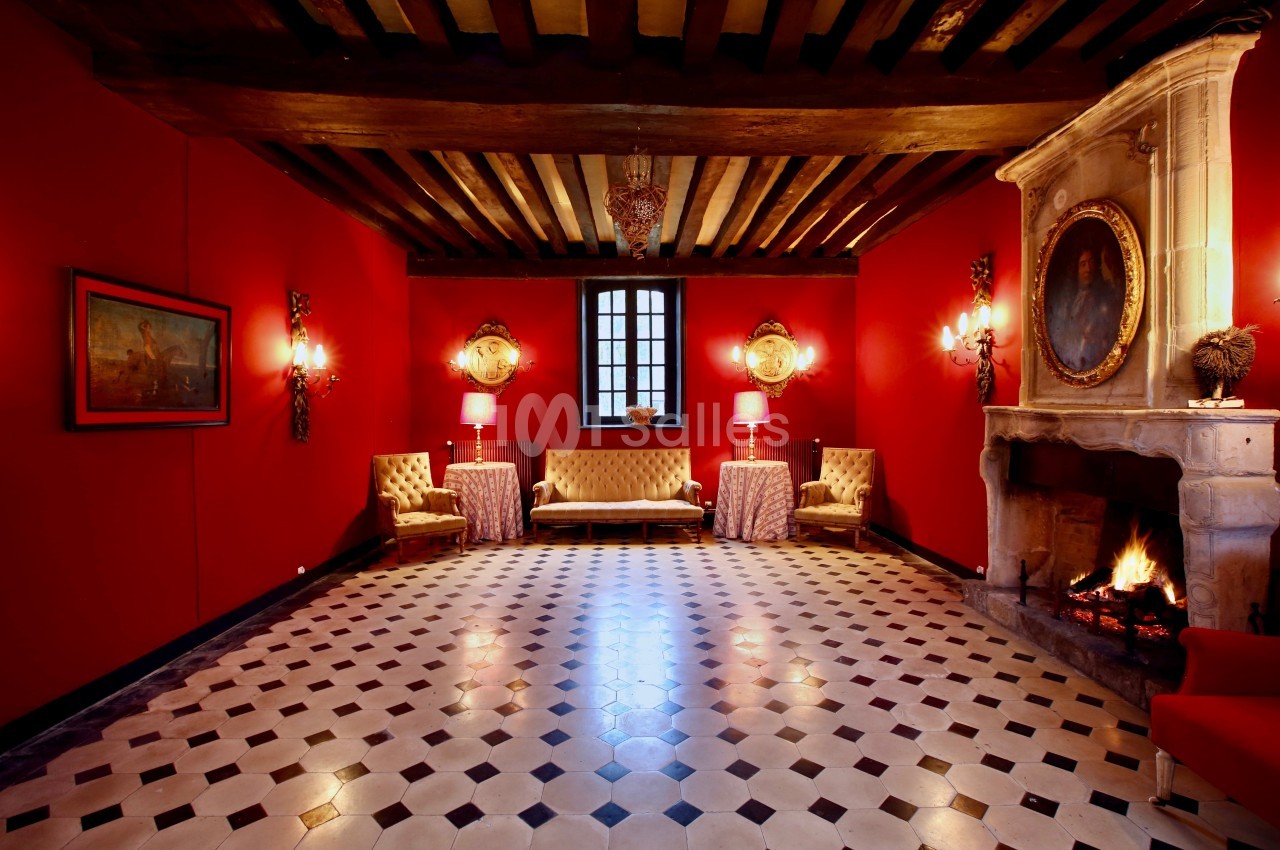 Salle avec murs rouges, cheminée allumée, plafond à poutres apparentes, mobilier classique et sol en damier.