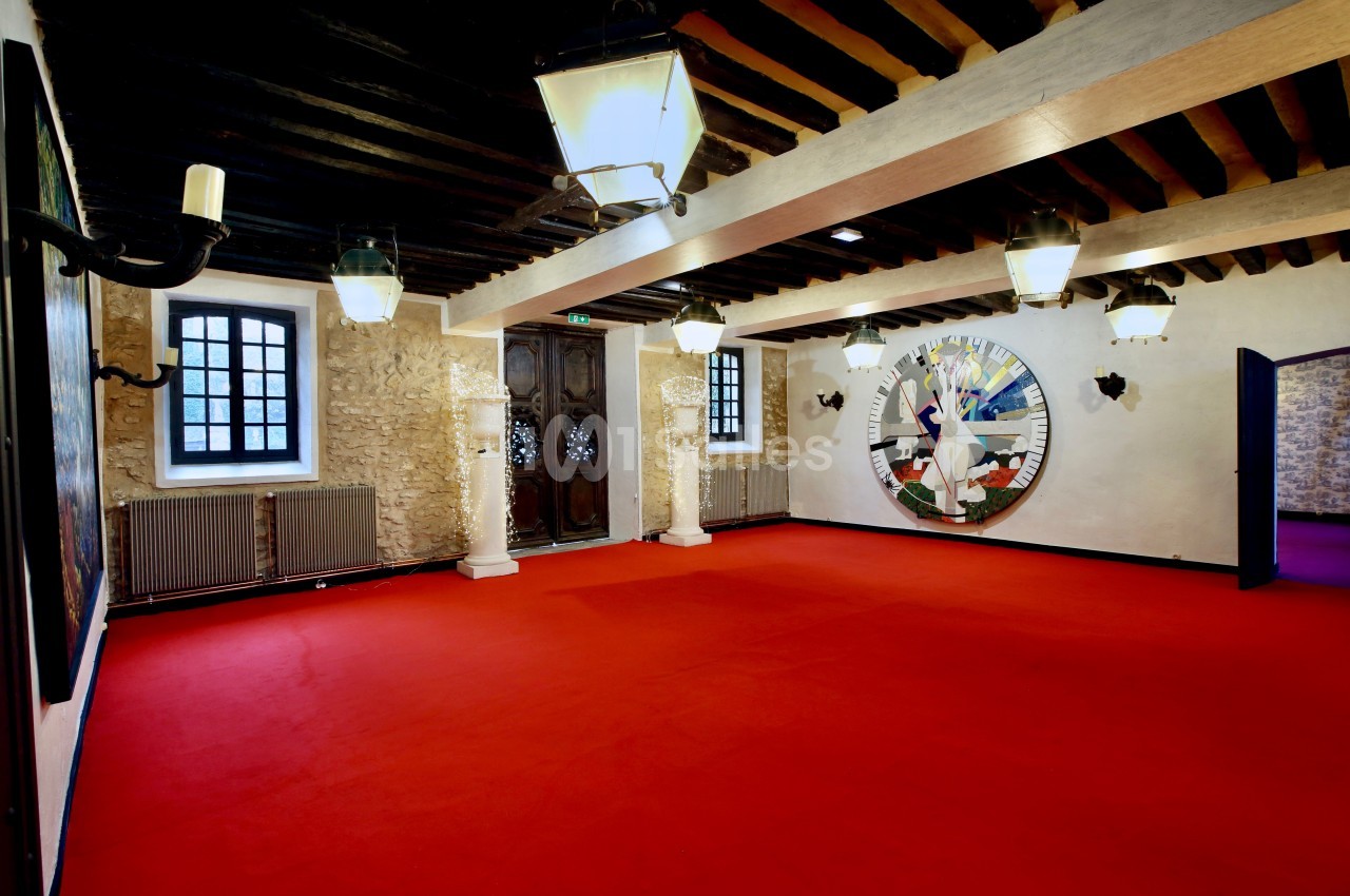 Salle avec un sol rouge, murs en pierre, poutres apparentes, luminaires suspendus et une grande horloge murale décorative.