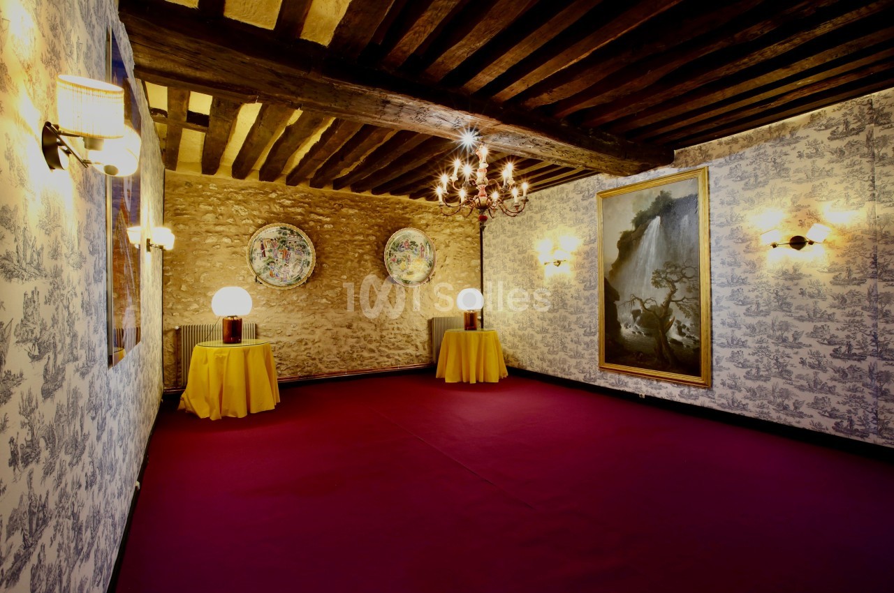 Salle avec plafond en poutres apparentes, murs en pierre et papier peint, tapis rouge et tables décorées de lampes.