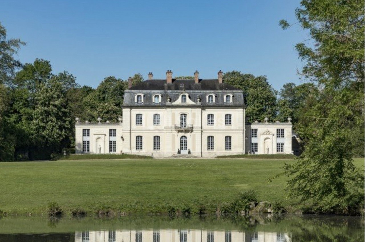 Façade d'un château blanc entouré de verdure, avec un étang au premier plan reflétant le bâtiment.
