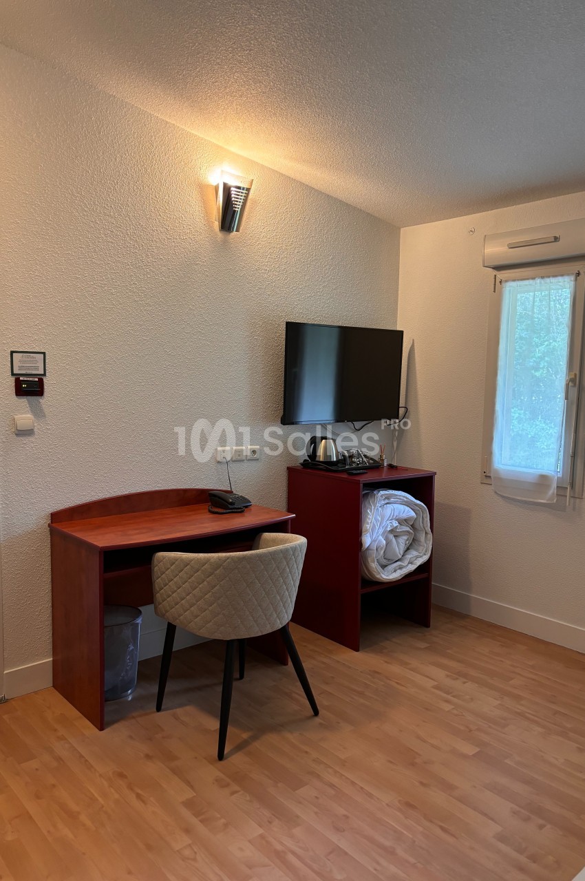 Bureau en bois avec chaise, télévision murale, étagère contenant une bouilloire et du linge, dans une pièce lumineuse.