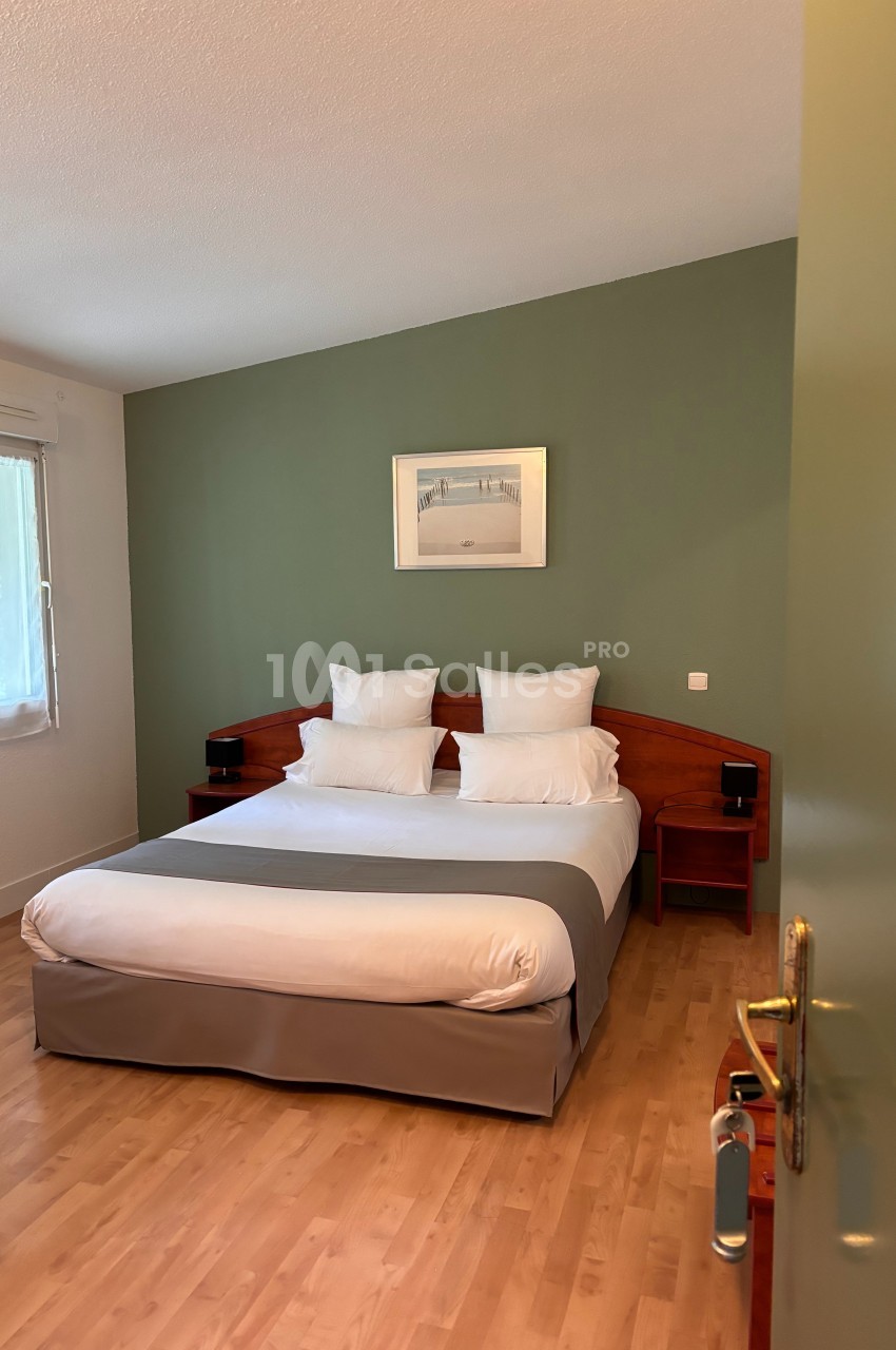Chambre avec lit double, draps blancs, mur vert, parquet clair, fenêtre avec rideaux et cadre décoratif au mur.