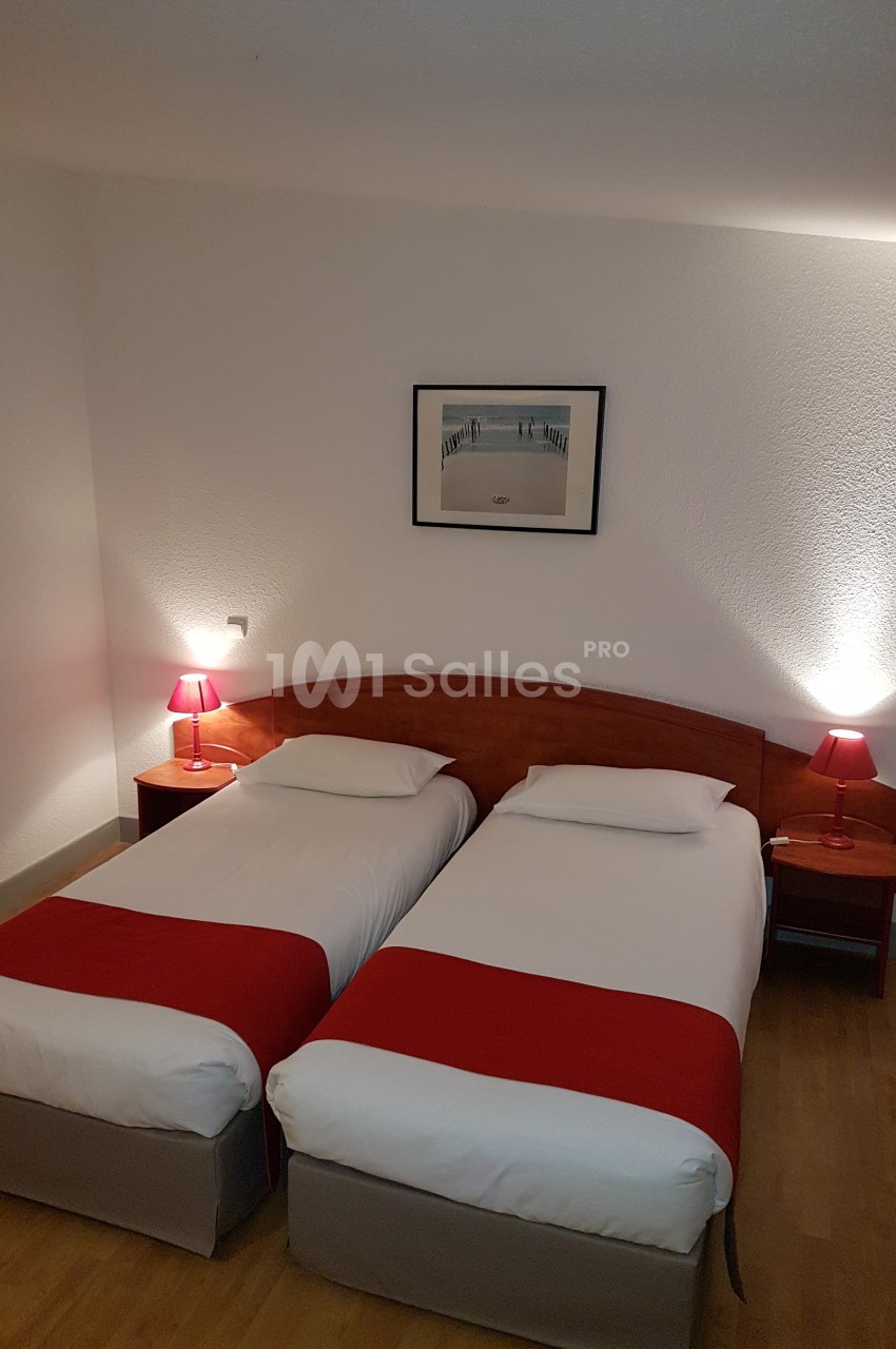 Deux lits simples avec draps blancs et chemins de lit rouges, encadrés par des lampes de chevet dans une chambre sobre.