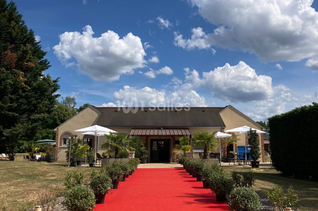 Façade d'un bâtiment avec une allée rouge bordée de plantes, entouré de parasols et de végétation sous un ciel bleu.